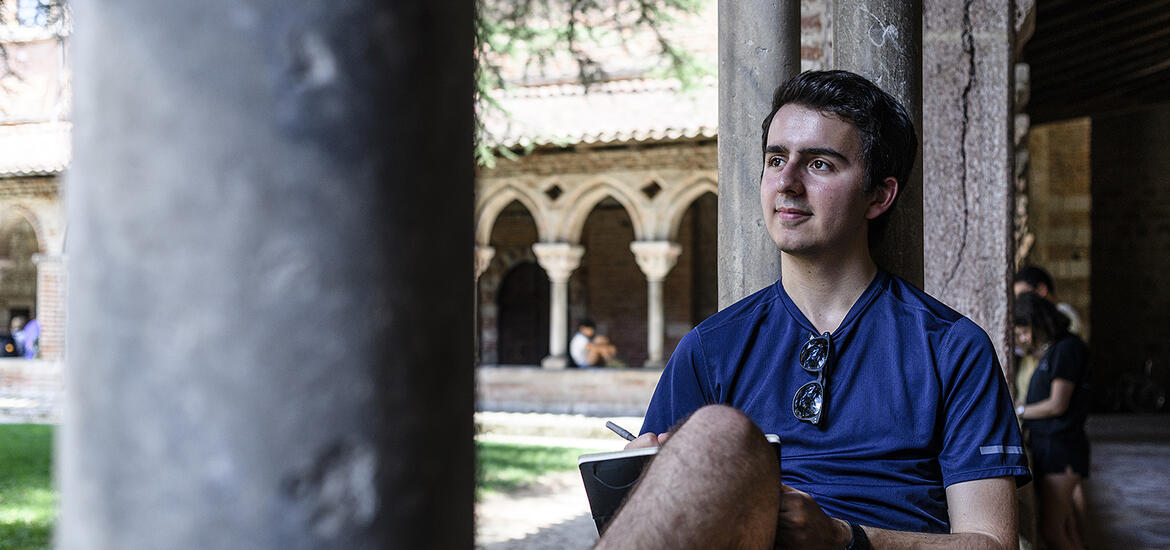A student sits outside in a cloister, leaning against a stone pillar with a notebook on one knee, in Moissac, France, during a pilgrimage