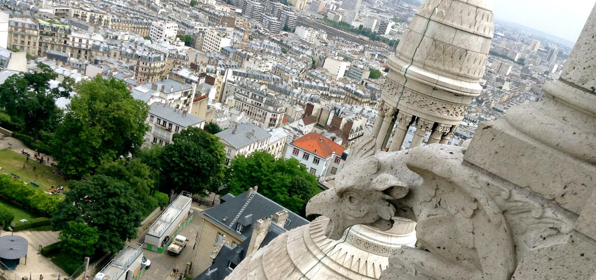 An aerial view of Paris from the Basilique du Sacré-Coeur, showing sprawling city buildings and streets, with a detailed stone gargoyle in the foreground