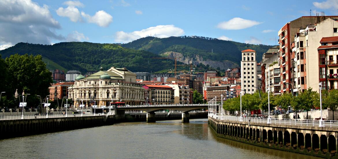 A view of La Ría river in Bilbao, Spain, lined with historic and modern buildings on both sides, a bridge, and green mountains in the background