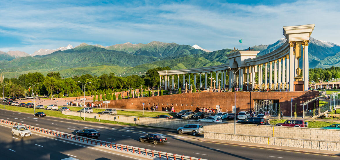A scenic view of Almaty, Kazakhstan, featuring a large colonnaded monument, a busy highway, and lush green trees, with snow-capped mountains in the background