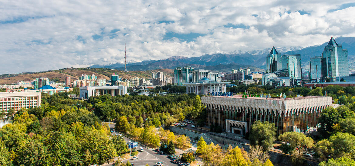  A panoramic view of Almaty, Kazakhstan, showing modern buildings surrounded by green spaces, with the snow-capped mountains in the background