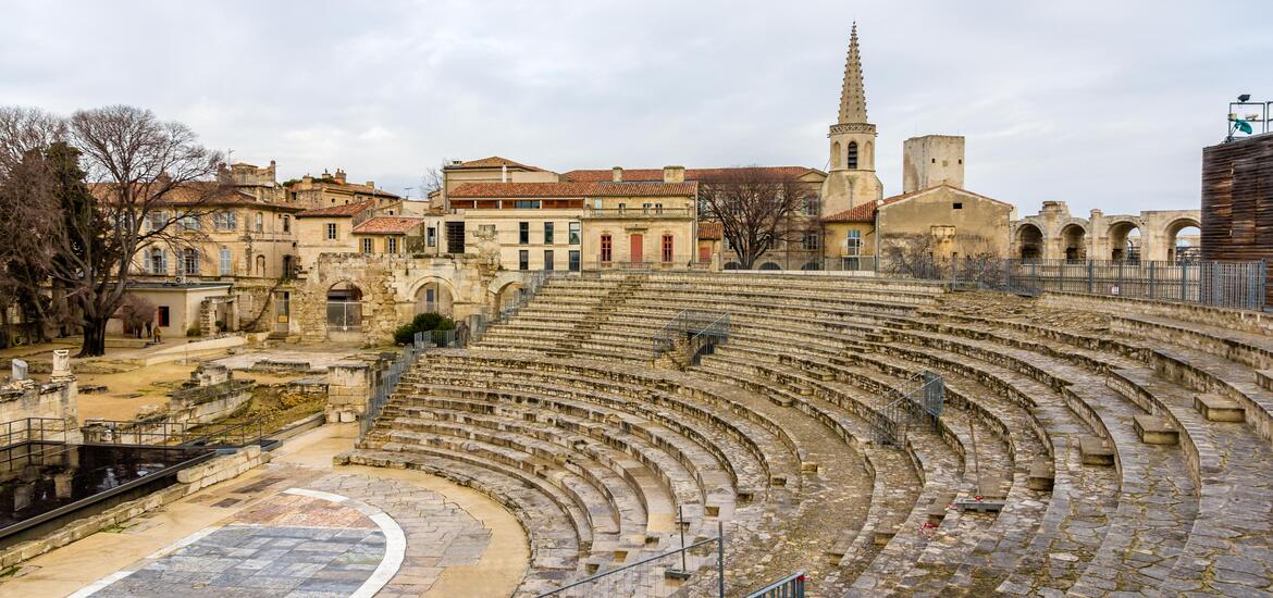 The stone ruins of the Roman Theatre in Arles, France, with tiered seating and historic buildings in the background, including a church with a pointed spire