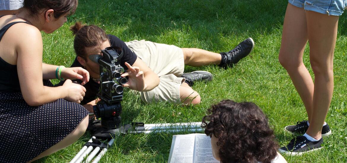 A female student points while looking at a large video camera held by a male student during a film project
