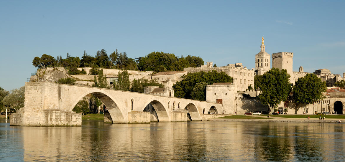 The historic Pont d'Avignon bridge stretches over the Rhone River with the medieval buildings of Avignon, including the Palais des Papes, visible in the background