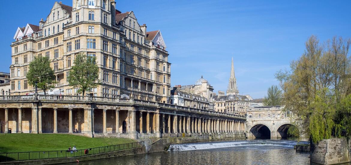 A view of historic buildings along the River Avon in Bath, Somerset, with classic stone architecture, trees, and a bridge