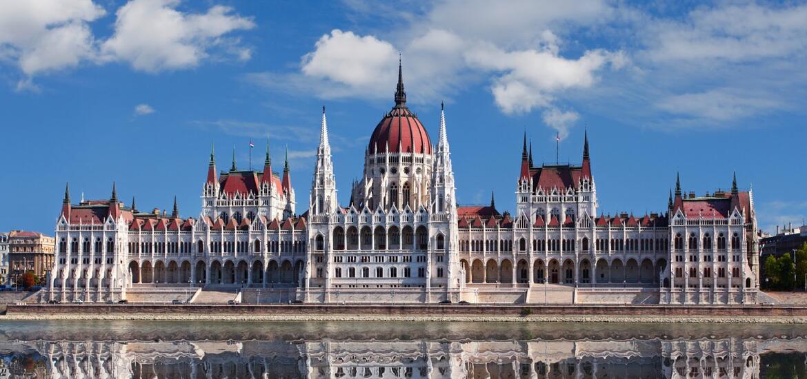 The iconic Hungarian Parliament Building with its red dome and spires, reflected in the waters of the Danube River under a bright blue sky