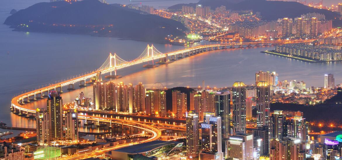 A panoramic view of Busan, South Korea at night, with the illuminated Gwangandaegyo Bridge spanning the water and tall buildings lighting up the city skyline