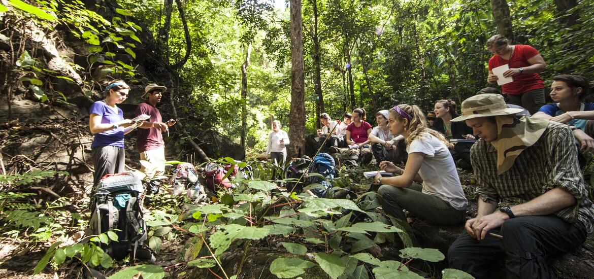 A group of students immersed in an outdoor learning session in the forest of Thailand, with notebooks and backpacks as they study 