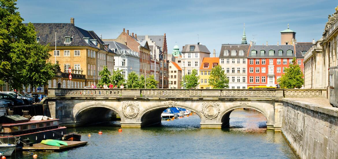 A picturesque view of a historic stone bridge over a canal in Copenhagen, lined with colorful, traditional buildings and trees