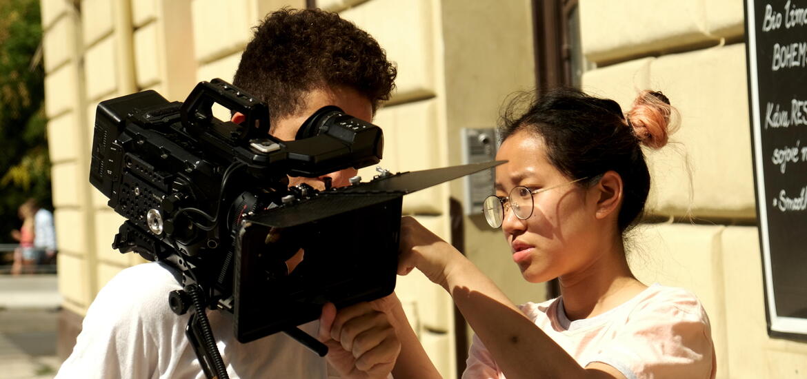 A female student points while looking at a large video camera held by a male student during a film project