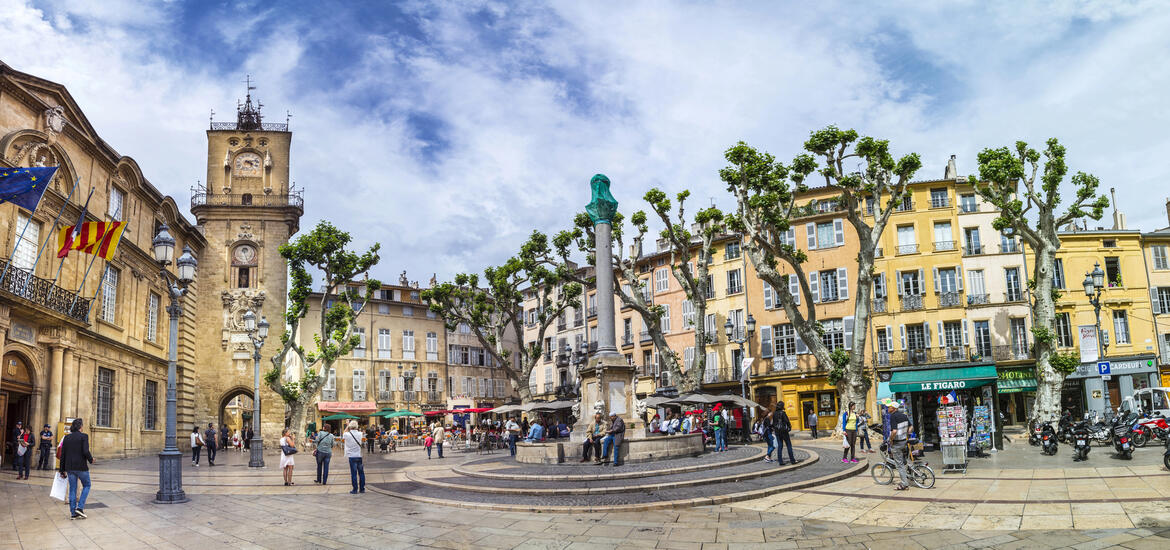 A bustling town square in Aix-en-Provence, France, featuring historic buildings with colorful facades, a clock tower, and a fountain, surrounded by leafy pruned trees