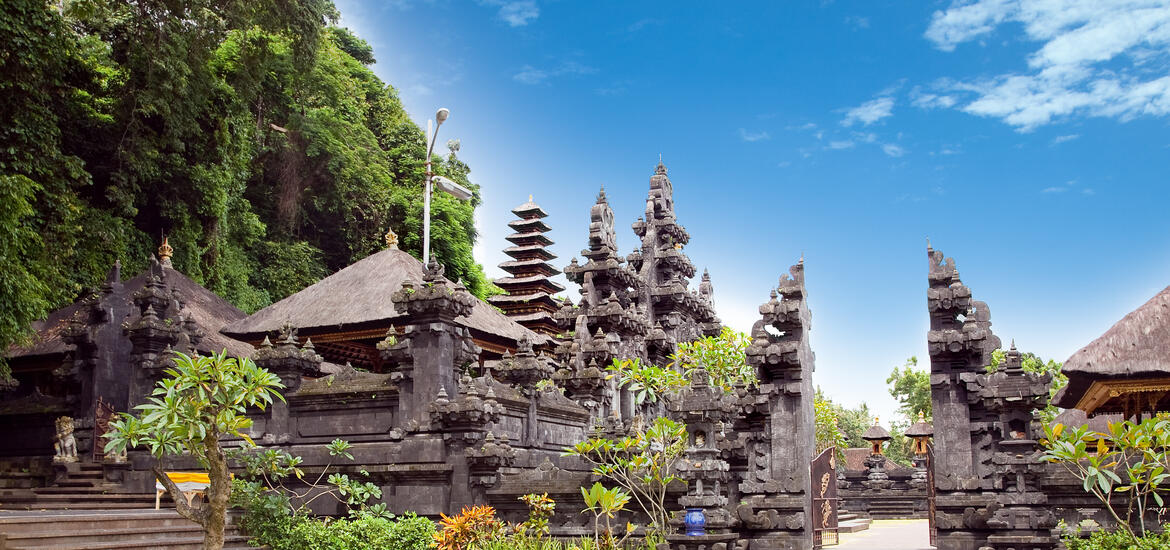 Pura Goa Lawah Temple, a traditional Balinese temple with ornate stone carvings, surrounded by lush greenery and a blue sky in Bali, Indonesia