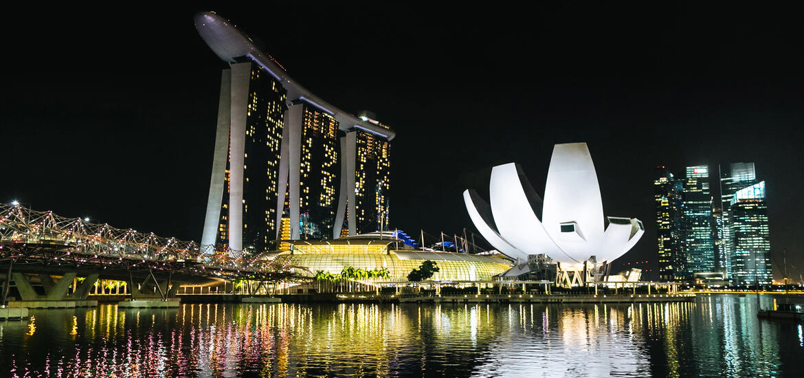The Singapore waterfront at night reflects colors on the bay from the lights of the helix bridge, SkyPark, the shoppes at Marina Bay Sands, and the lotus shaped ArtScience Museum