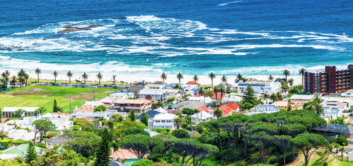 A coastal view of Cape Town, South Africa, featuring beachfront homes, lush greenery, and palm trees along the shoreline, with turquoise water and waves