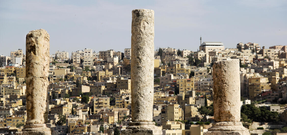 Ancient stone columns from the Roman-era ruins stand in the foreground, with the cityscape of Amman in the background