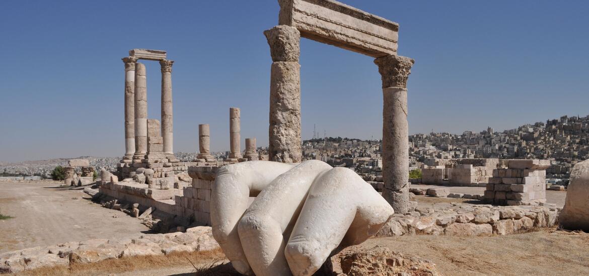 The ancient ruins of the Hercules Temple in Amman, Jordan, featuring partially standing columns and the large stone 'Hand of Hercules' sculpture, with the cityscape in the background