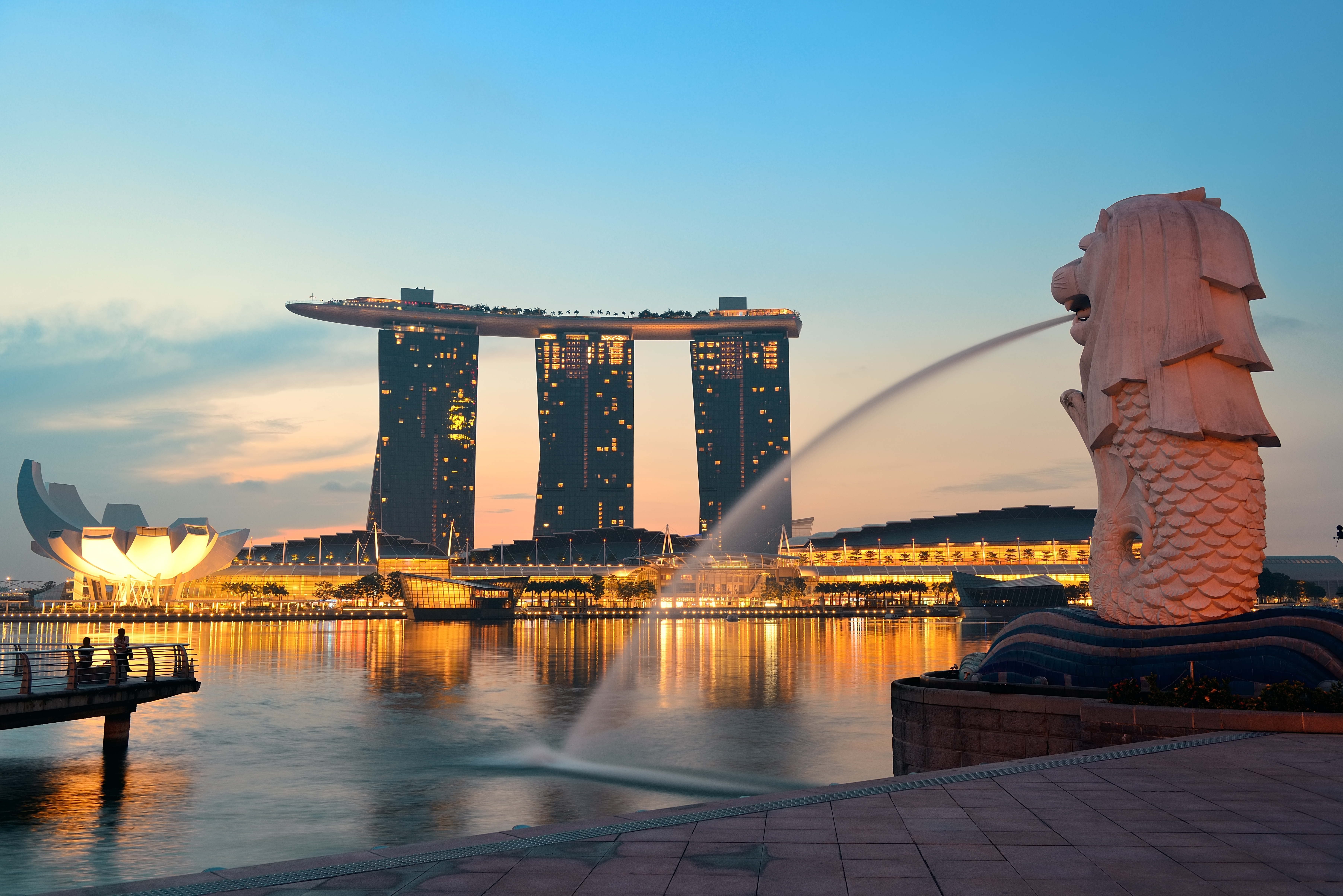 A view of Singapore's iconic Merlion statue with water spouting from its mouth, overlooking the Marina Bay Sands hotel and skyline at dusk, with the bay reflecting the evening lights.