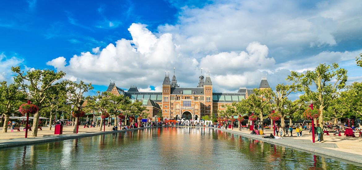 The Rijksmuseum in Amsterdam viewed from Museum Square, with a reflecting pool, trees, and and people walking on a sunny day