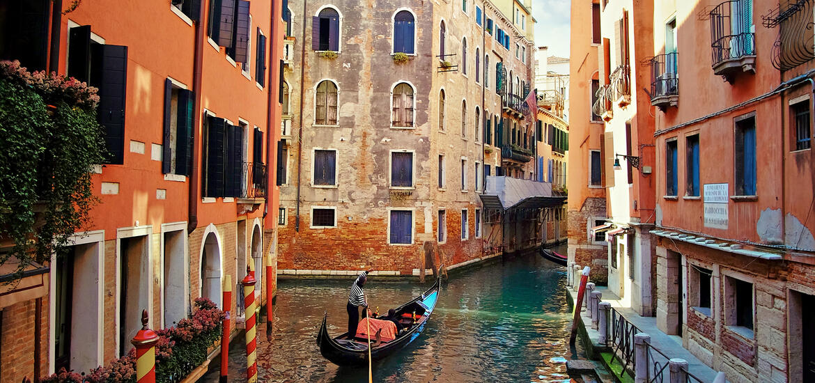 A gondola glides through a narrow canal in Venice, surrounded by vibrant orange and weathered brick buildings with arched windows and balconies