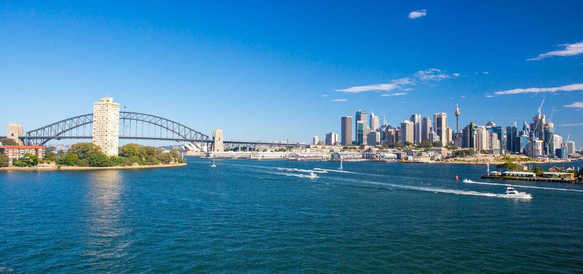 Panoramic view of Sydney Harbour, featuring the iconic Harbour Bridge and the city's skyline under a clear blue sky, with boats moving across the water