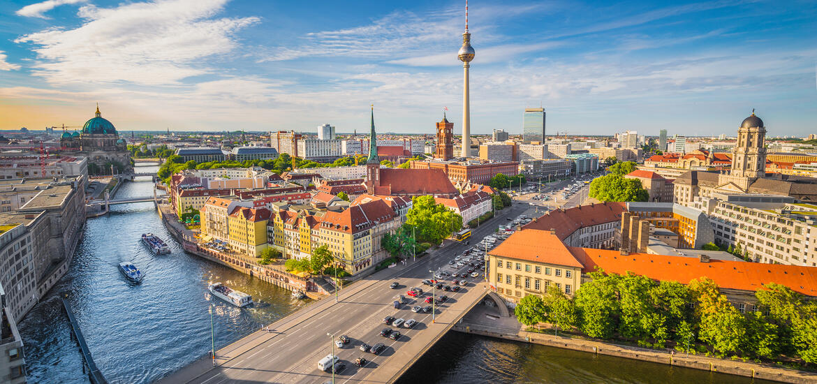 A panoramic view of a city skyline, with a river running on the lefthand side and buildings with red roofs on its bank
