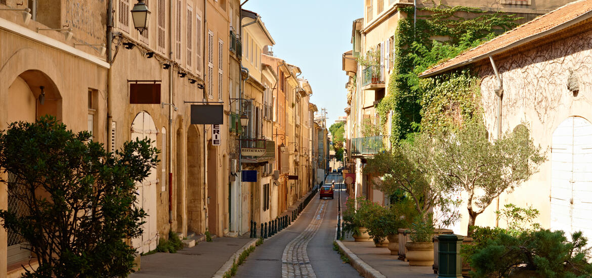 A narrow, sunlit street in Aix-en-Provence, France, lined with charming buildings, shutters, and potted plants, leading into the distance 