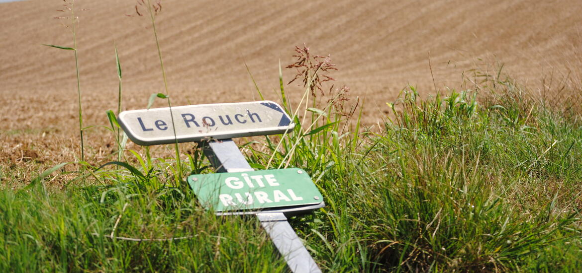 Two signs in French on a signpost, one reading 'Le Rouch' and the other 'Gîte Rural,' leaning in tall grass with a brown plowed field in the background
