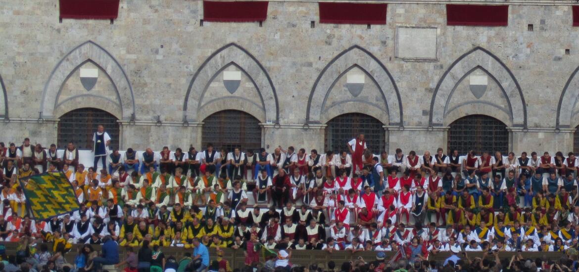 A large group of people dressed in colorful traditional attire seated in front of a stone building with arched windows, preparing for the Palio di Siena, a historic horse race held in Siena, Italy