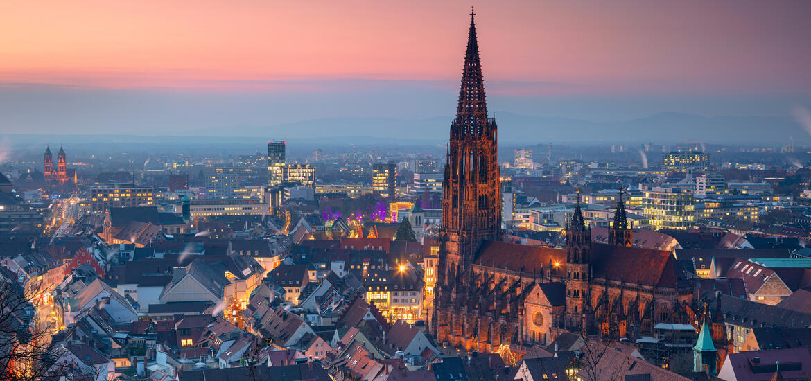 A panoramic view of Freiburg, Germany, at twilight, featuring the illuminated spire of the Freiburg Minster and the softly glowing city skyline 