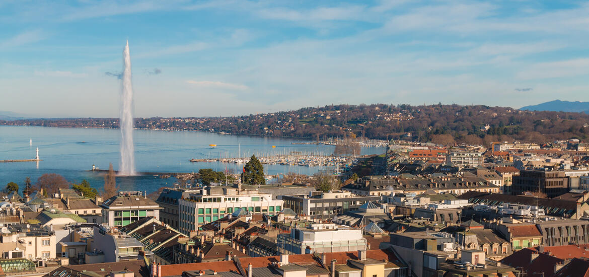 An aerial view of Geneva, Switzerland, featuring the Jet d’Eau fountain shooting water high above Lake Geneva, with the city rooftops in the foreground 