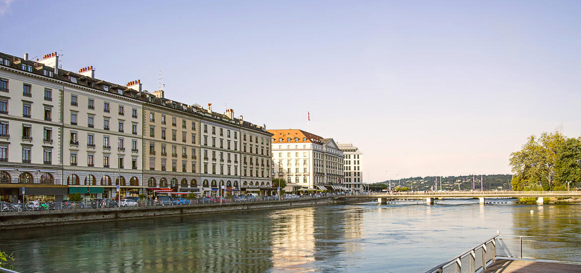 A view of the Geneva Lake embankment in summer, featuring historic multi-story buildings along the waterfront with a bridge in the distance