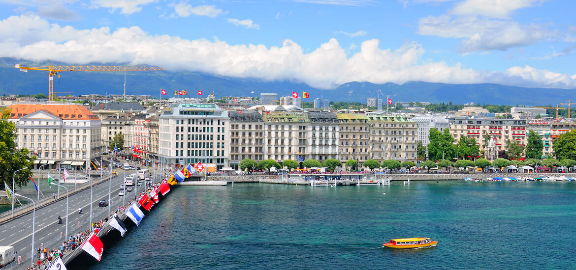 A panoramic view of Geneva, Switzerland, featuring the lakeside buildings, a bridge with colorful flags, and mountains in the background