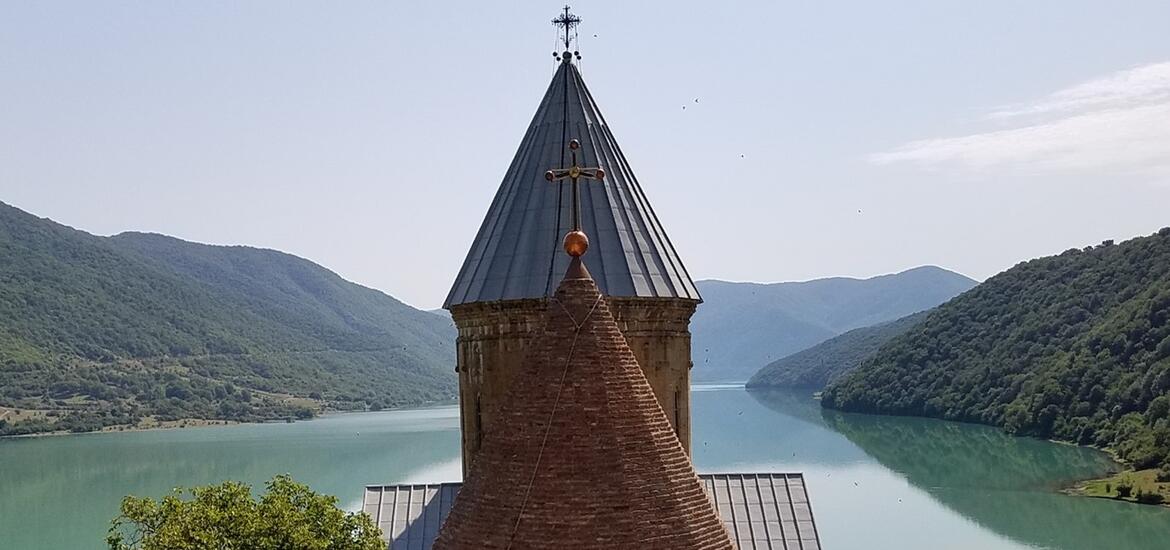 The pointed towers of Ananuri Castle rise in front of a serene river and surrounding green mountains