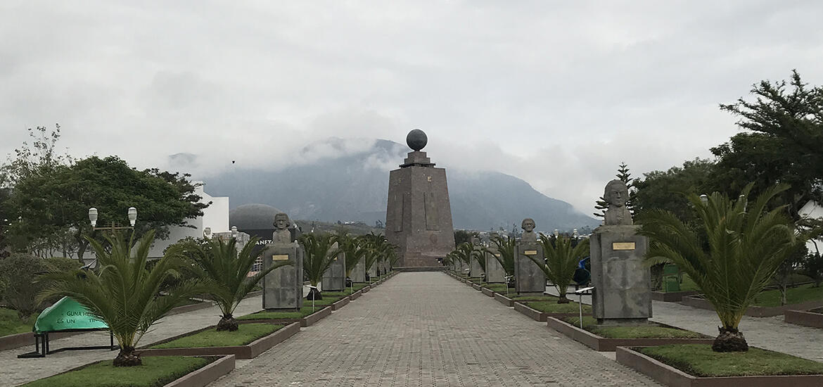 A tree-lined boulevard leads to the Mitad del Mundo monument in Ecuador, a large stone structure marking the equatorial line, with misty mountains in the background