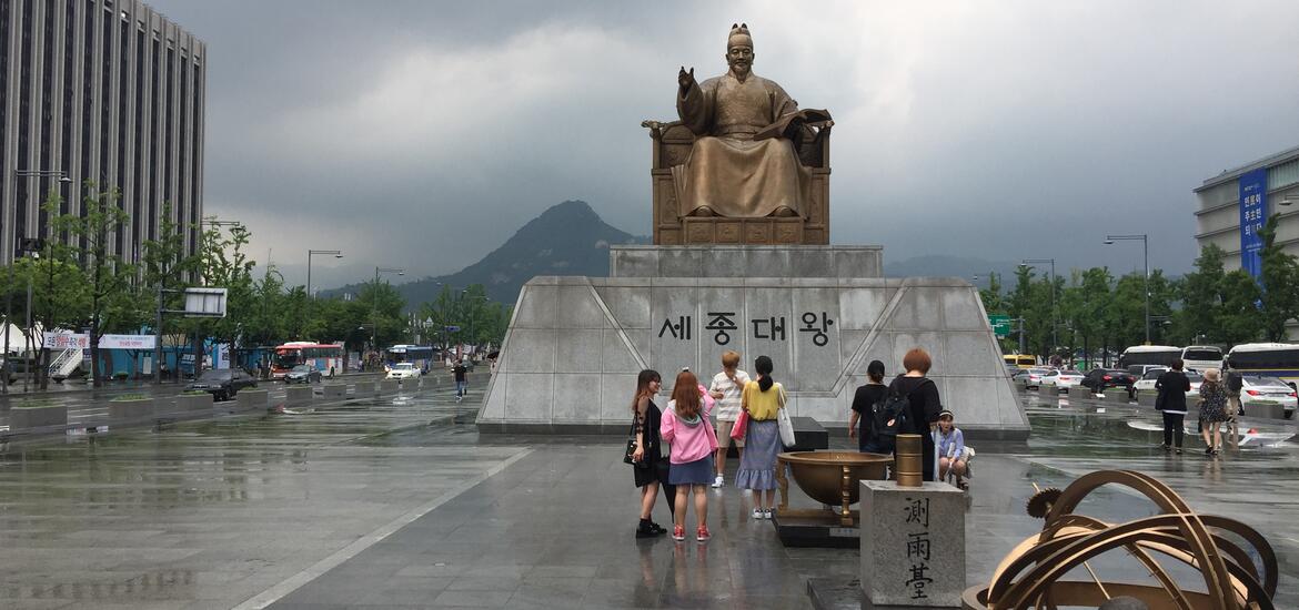 A view of the King Sejong statue in Gwanghwamun Plaza, with people gathered around the monument under a cloudy sky, and mountains visible in the background