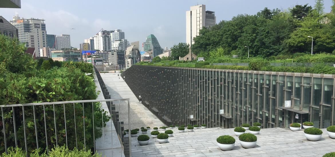 A view of Ewha University in Seoul, featuring a unique, sunken pathway lined with glass buildings, surrounded by greenery and urban high-rise buildings in the background