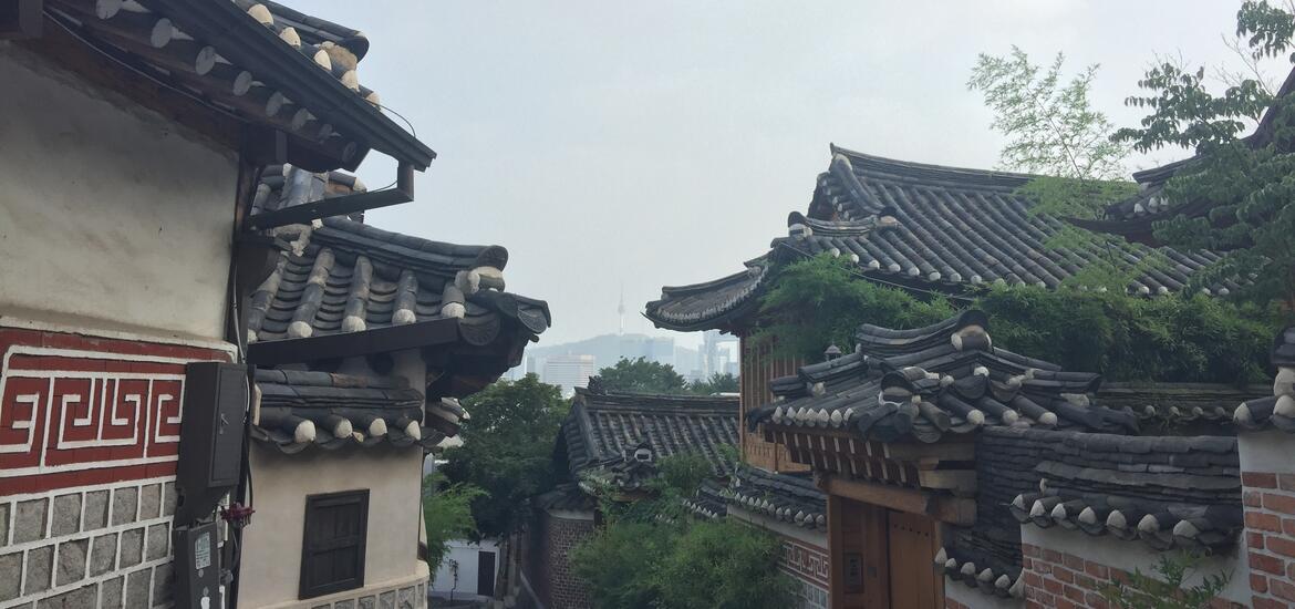 A view of Bukchon Hanok Village in Seoul, featuring traditional Korean tiled rooftops with Namsan Tower faintly visible in the hazy background