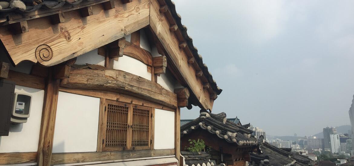 A close-up view of a traditional wooden roof and window details in Bukchon Hanok Village, Seoul, with additional tiled rooftops and the city skyline visible in the background