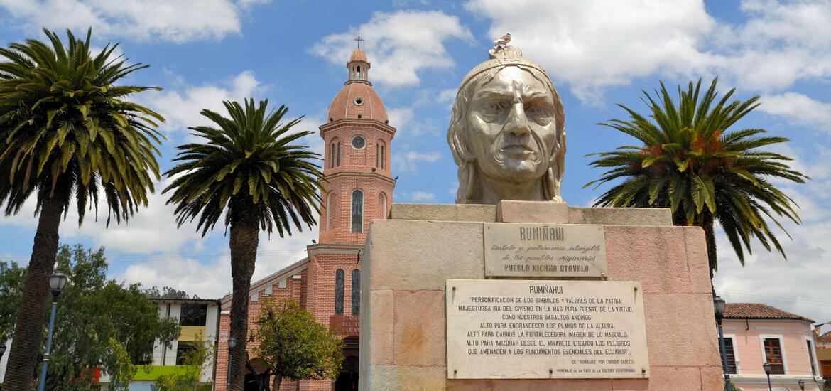 A tree-lined boulevard leads to the Mitad del Mundo monument in Ecuador, a large stone structure marking the equatorial line, with misty mountains in the background