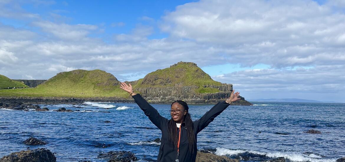 Jasmine McKenzie stands smiling with arms raised in front of the rugged cliffs and green hills of Giant's Causeway