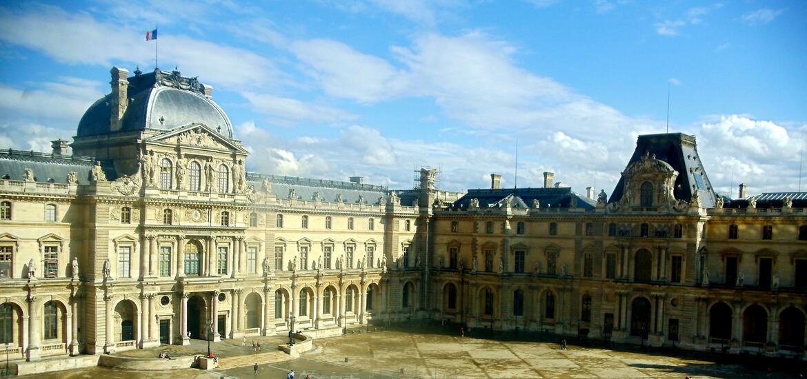 The Louvre Museum, a grand and ornate complex of stone buildings with intricate facades, bathed in sunlight under a clear blue sky