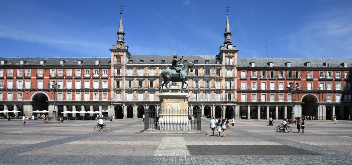 A view of Madrid's Plaza Mayor, featuring the statue of King Philip III at the center, surrounded by red buildings with arched entrances and spired towers