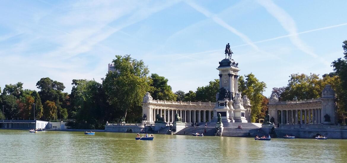 A view of the pond in Buen Retiro Park, Madrid, with small boats on the water and the grand monument to Alfonso XII surrounded by greenery