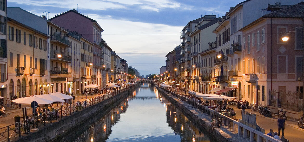 A twilight view of The Navigli district in Milan, with bustling outdoor cafes and restaurants lining the canal, illuminated by streetlights