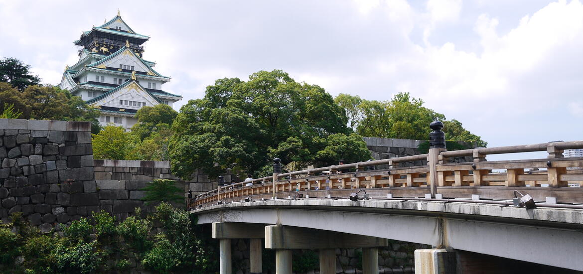 A view of Osaka Castle in Japan, with its multi-tiered roof and ornate details, seen from across a wooden bridge surrounded by stone walls and greenery