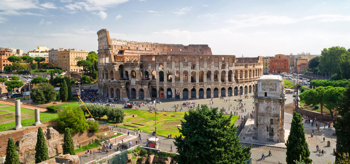 An aerial view of the Colosseum in Rome, Italy, captured from Palatine Hill, showing the ancient amphitheater surrounded by green spaces, nearby buildings, and visitors exploring the site