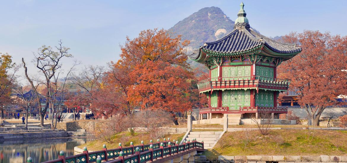 A view of Gyeongbokgung Palace in Seoul, featuring a traditional pavilion surrounded by autumn foliage and set against a mountain backdrop