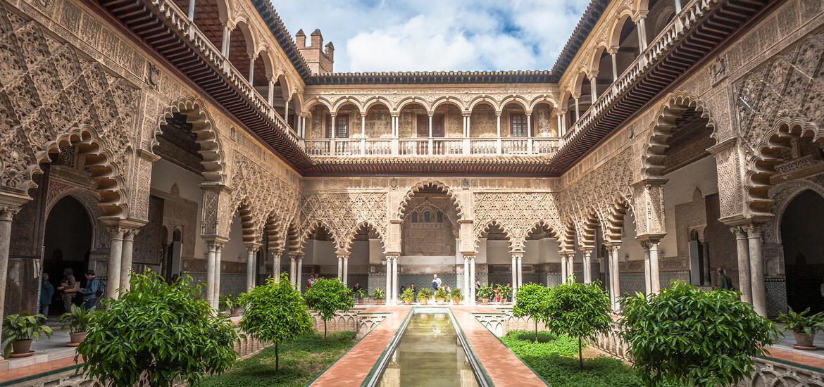 Moorish architecture in the courtyard of the Alcázar of Seville, with arched doorways, detailed carvings, and greenery surrounding a narrow reflecting pool