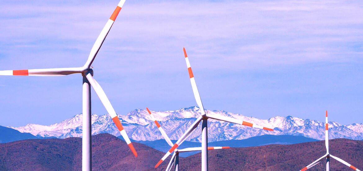 Wind turbines spin in the foreground with snow-capped Andes mountains in the background, under a clear sky in Northern Chile