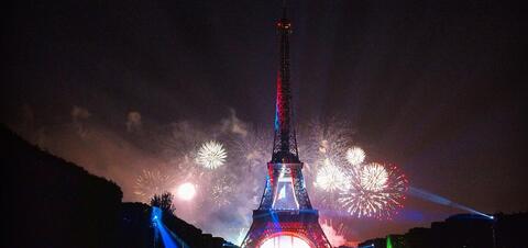 The Eiffel Tower illuminated with blue and red lights, surrounded by vibrant fireworks lighting up the night sky in Paris, France
